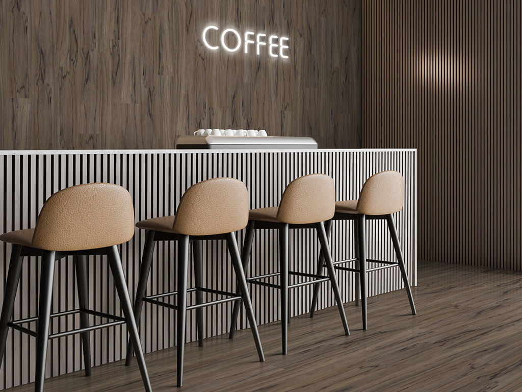 A coffee bar setting, with brown woodgrain luxury vinyl tile adorning its walls and floor. A white neon light spells out the word "coffee." Four camel-colored bar stools with dark metal legs sit in front of a coffee bar with vertical slats.