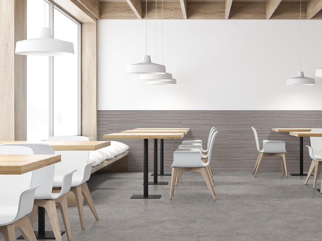 A corner of a cafe, decorated with minimalist design elements, muted and neutral tones, with light oak colored tables, each with a single black metal leg, white Scandinavian-style chairs with light oak legs, and white pendant lights hanging above the tables. The back wall is decorated with grey luxury vinyl tile on the bottom half, while the top half is white. The cafe features concrete-look luxury vinyl tile flooring. Light oak wood beams are exposed across the ceiling.