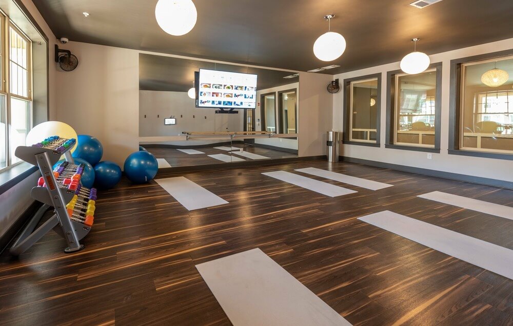 A multifamily housing complex fitness room, featuring wood look LVT flooring, light grey yoga mats, a mirrored wall, and various dumbbell and exercise ball equipment.