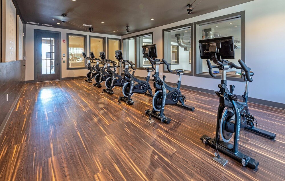 A multifamily housing complex fitness room, featuring wood look LVT flooring, six stationary workout bikes and window-filled walls.