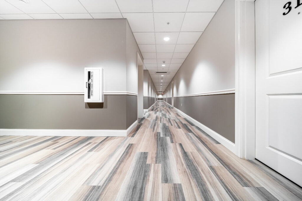 A multifamily housing complex shared hallway, featuring dual-toned white and grey walls and wood look LVT with a blend of beige, grey and light brown undertones.