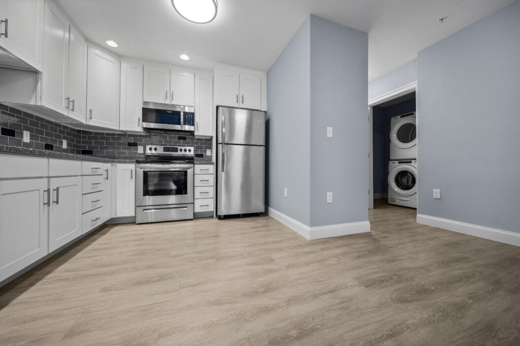 A kitchen area, featuring wood look LVT with light grey undertones, bluish grey walls, white cabinets, and a dark grey tile backsplash.