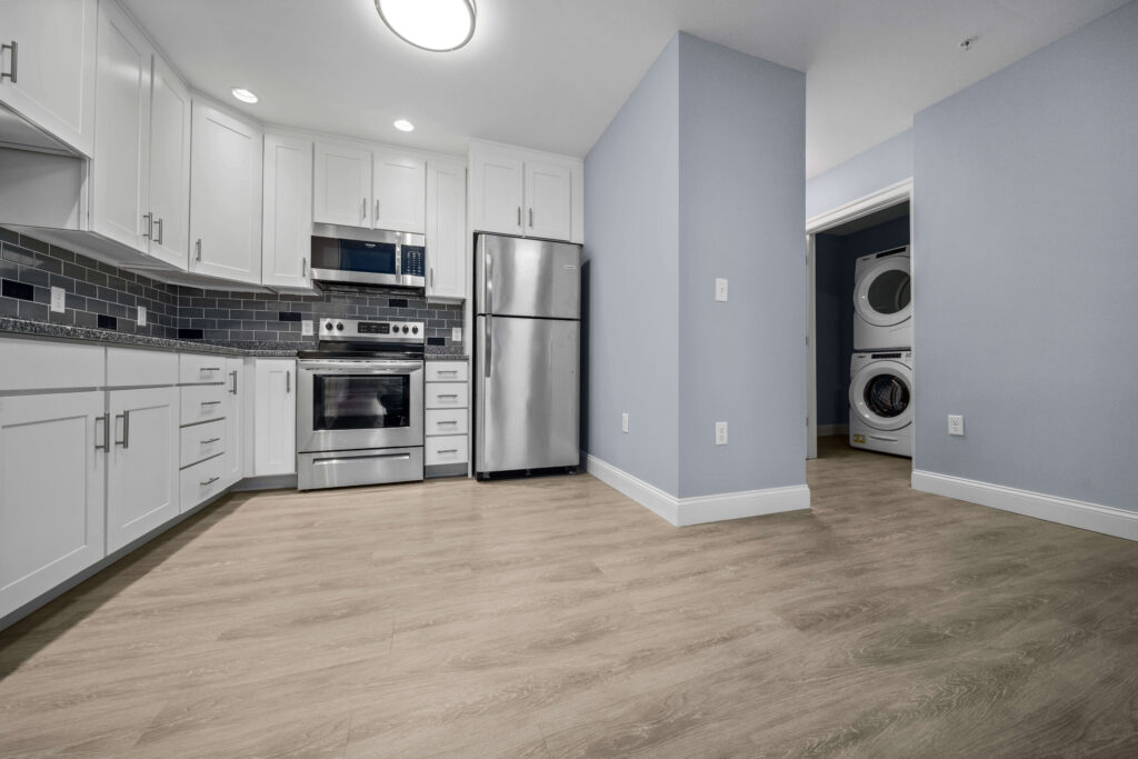 Modern kitchen with white cabinetry, stainless steel appliances, and Sharkskin LVT flooring. On the right is a laundry nook with a stacked washer and dryer.