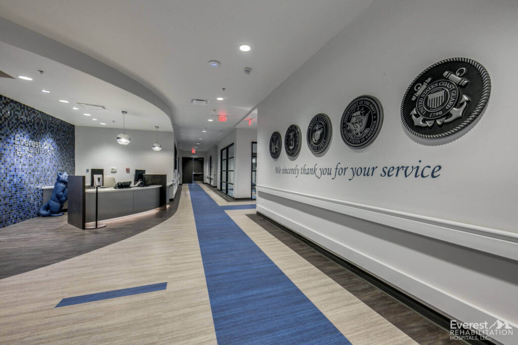  Welcoming corridor with a tribute to military service, featuring a sleek reception desk, blue and grey color palette, and LVT flooring, integrating modern healthcare design elements.