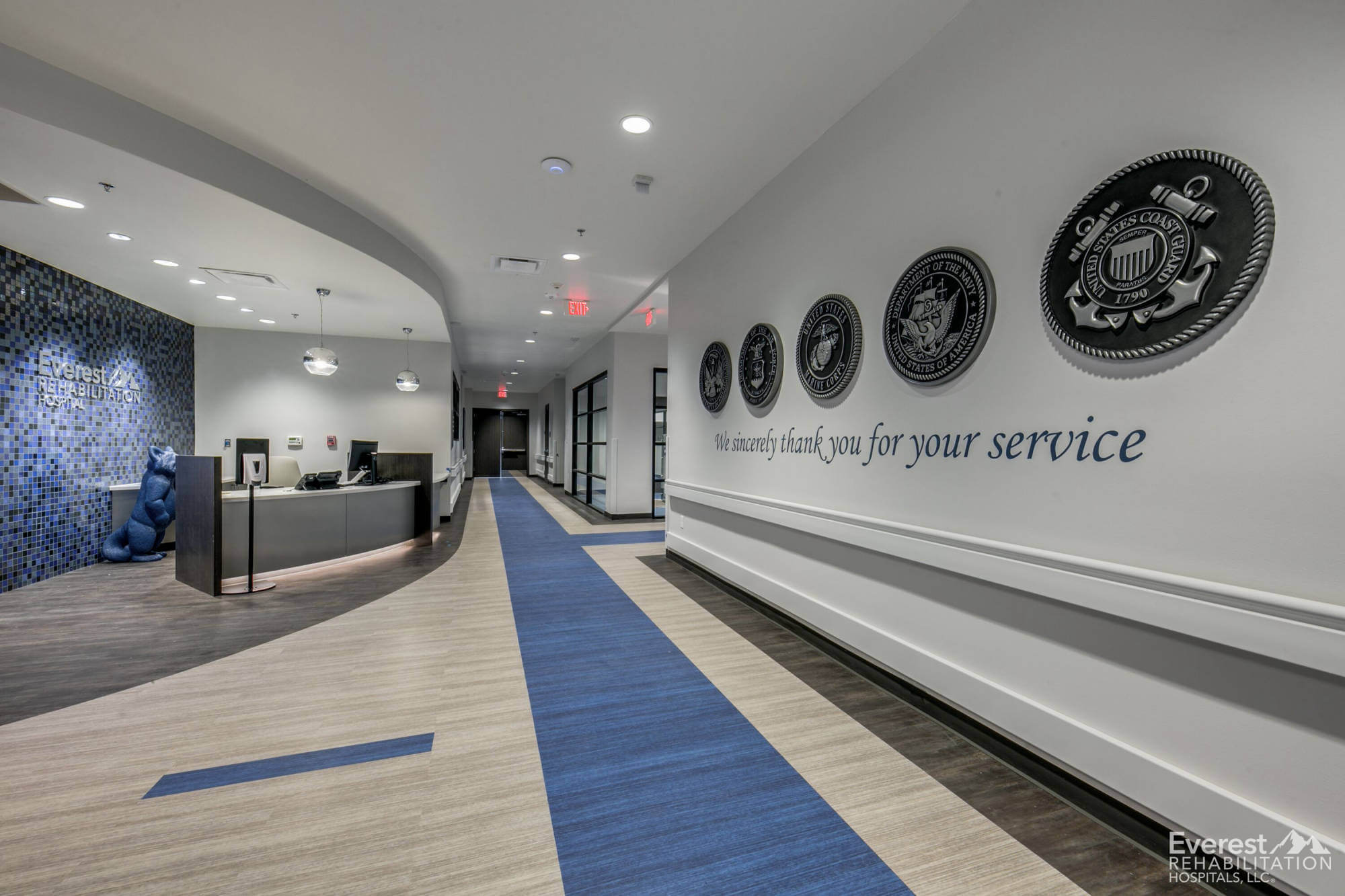Welcoming corridor with a tribute to military service, featuring a sleek reception desk, blue and grey color palette, and LVT flooring, integrating modern healthcare design elements.