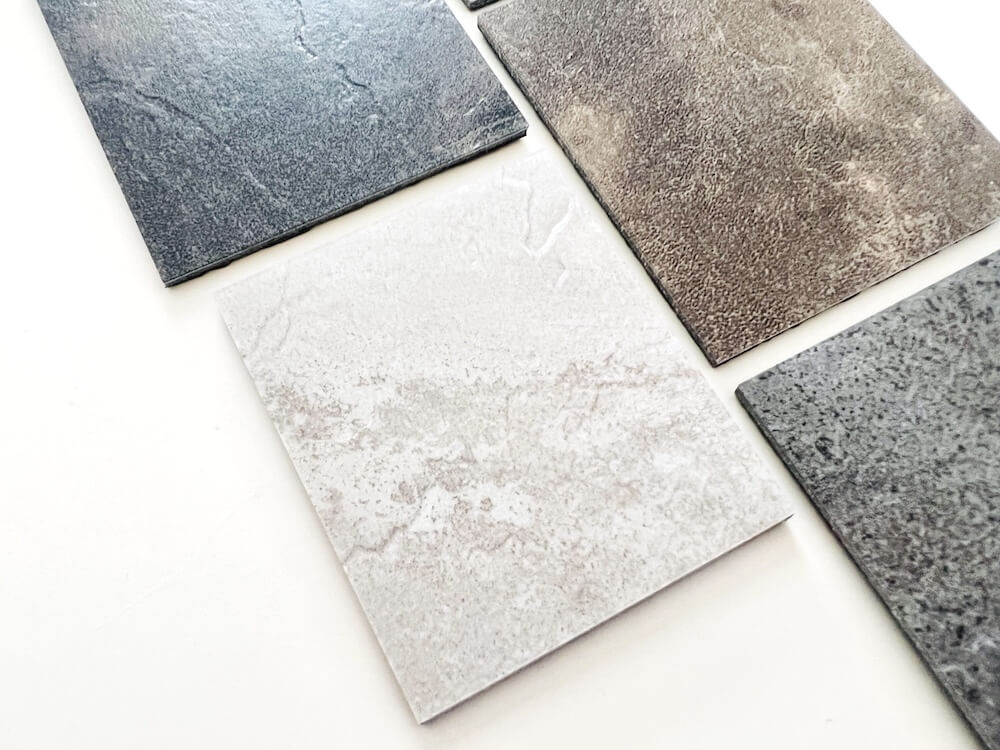 The image showcases an array of luxury vinyl tile (LVT) samples, presenting a variety of textures and colors that mimic the appearance of polished or stained concrete. The tiles display different finishes, ranging from subtle marbling to more pronounced textured surfaces, in shades of gray, taupe, and off-white. This selection highlights LVT’s ability to offer a clean, consistent aesthetic while capturing the trend of concrete’s visual appeal, without the imperfections like existing stains or adhesive marks that natural concrete might exhibit.