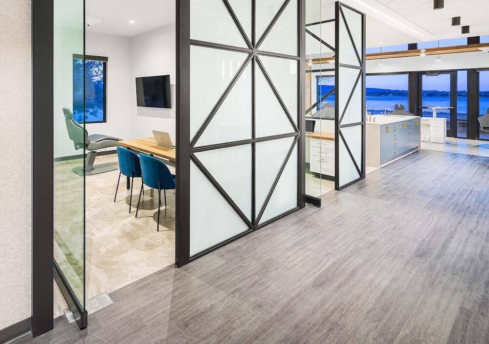 The image features a modern dentist office with a sophisticated interior design. It showcases a seamless transition between different flooring materials—luxury vinyl tile (LVT) that imitates a light gray wood texture in the main area and a polished concrete look in the private office space. The office is equipped with contemporary furniture and vibrant blue chairs that provide a pop of color. Large glass partitions with black metal frames divide the spaces, enhancing the open and airy feel of the office while offering privacy where needed. This setting demonstrates the versatility of LVT in creating an elegant and functional environment suitable for professional spaces like a dentist's office.