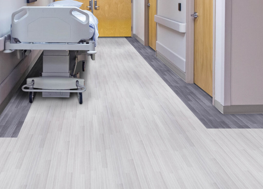 A healthcare facility’s hallway with seamless light gray wood-look vinyl sheet flooring, a hospital bed, and contrasting gray trim for navigation and design.