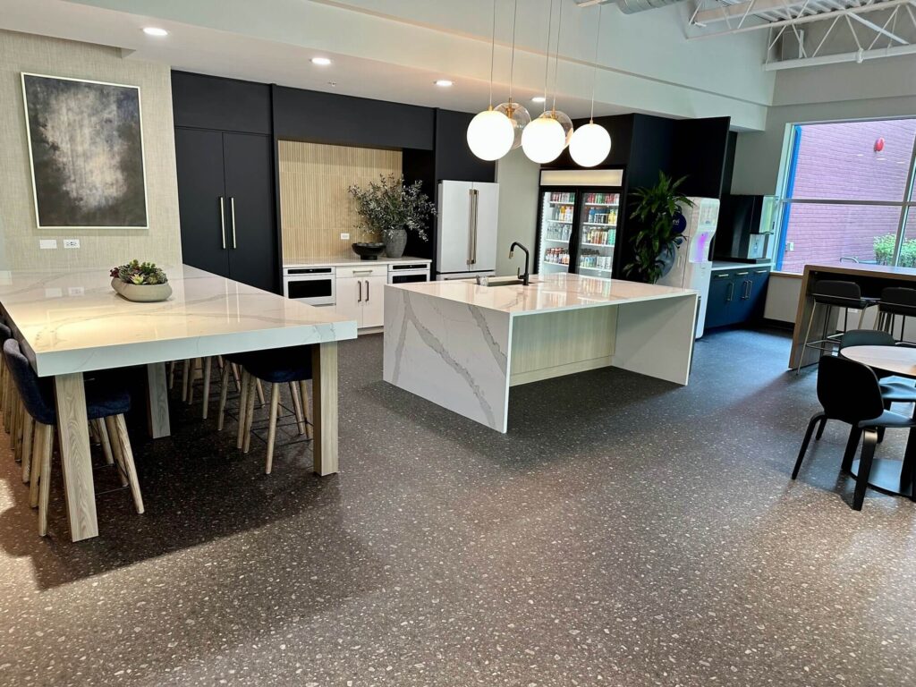 A commercial kitchen with dark terrazzo-look LVT flooring, a white marble island, contemporary lighting, and a dining area with matching chairs and a refrigerator in the background.]